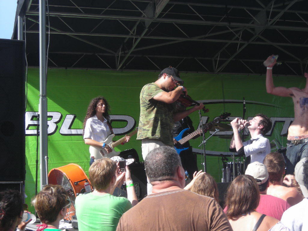 Eine Gruppe von Musikern, die verschiedene Instrumente auf einer Bühne mit einem großen schwarzen Lautsprecher links spielen, während das Publikum davor steht und die Vorstellung genießt, mit einem grünen Banner im Hintergrund.