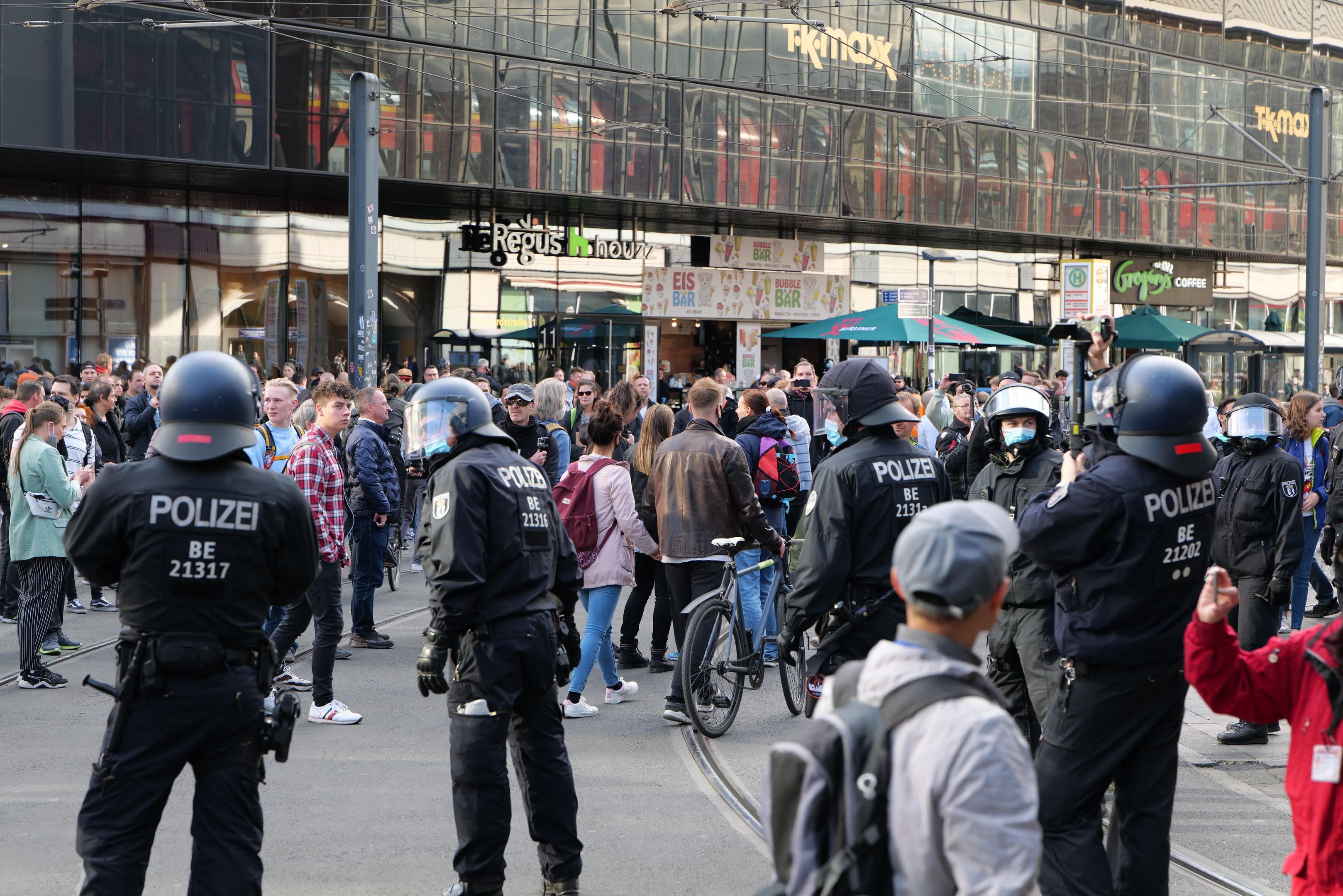 Polizeibeamte vor einer Menschenmenge auf der Straße stehend, einige halten Fahrräder und tragen Helme, mit Gebäuden und Schirmen im Hintergrund.