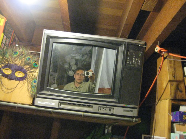 Ein Fernsehbildschirm, der eine Person umgeben von Pfauenfedern, Büchern und Schachteln zeigt.