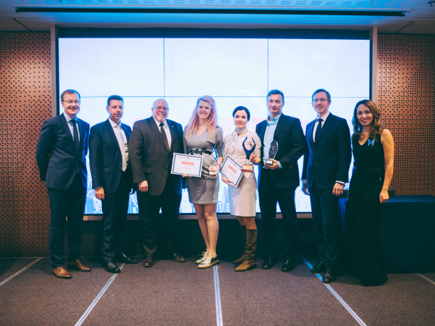 Eine Gruppe strahlender Menschen hält Zertifikate und steht vor einem Bildschirm, rechts davon ein auf einem schwarzen Tuch gedeckter Tisch und ein Gegenstand an der Wand im Hintergrund, feiert ihren Sieg bei den International Business Awards 2018.