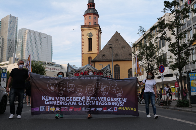 Eine Gruppe von Menschen mit Masken, die eine Banner mit Text und Bildern tragen, geht eine Straße entlang, mit einem geparkten Auto auf der linken Seite, Gebäude, Bäumen, Schildern, Masten, einem Uhrenturm und einem klaren blauen Himmel im Hintergrund.