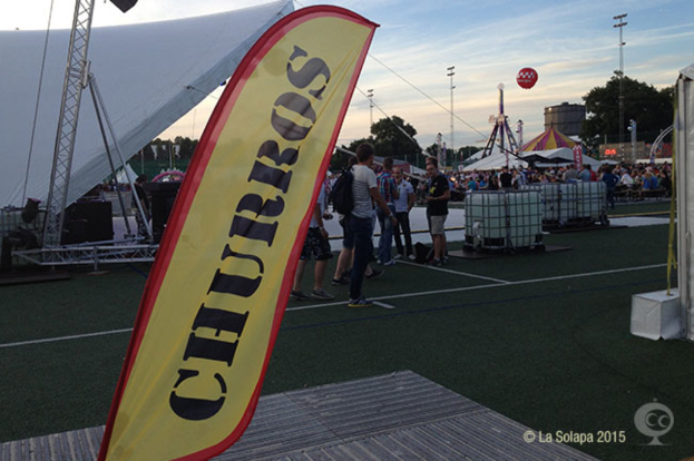 Eine Karnevals Szenerie mit Zelten, einem zentralen Mast, Menschen, Bäumen, Strommästen, Straßenbahnen auf der Straße und einem eisernen Ständer auf der linken Seite unter einem Himmel-Hintergrund.
