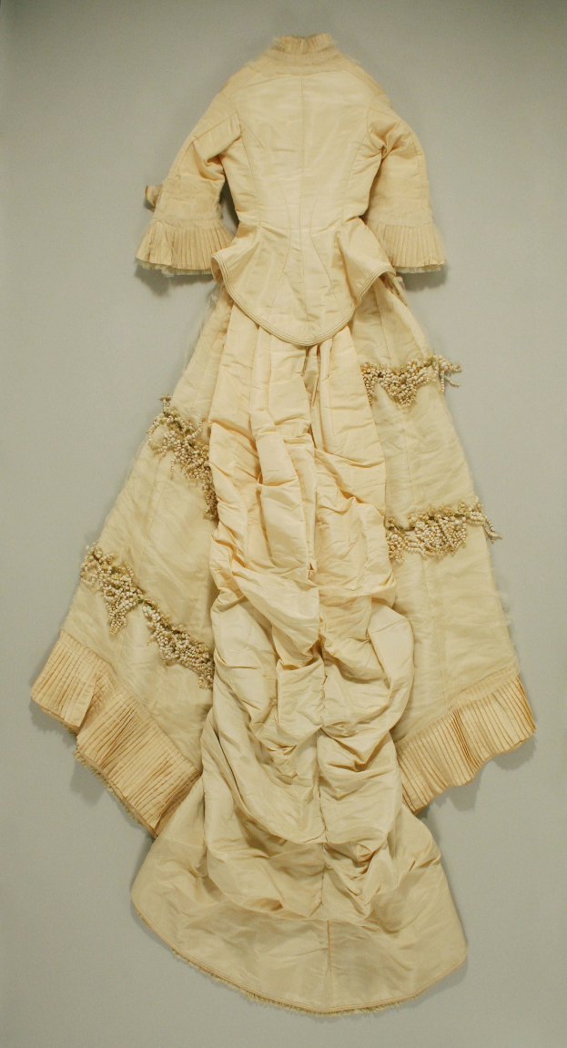 Ein cremefarbenes, bodenlanges Kleid mit engem Oberteil, langen Ärmeln und hohem Kragen, verziert mit feiner Stickerei und Pailletten, präsentiert in einem Museum mit einem üppigen, gerafften Rock.