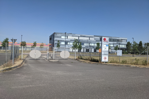 Ein großes Gebäude mit vielen Fenstern, identifiziert als First Solar, ist von einem Metallzaun, Bäumen, Pflanzen, Gras, Schildern, Straßenlaternen und einem Weg umgeben.