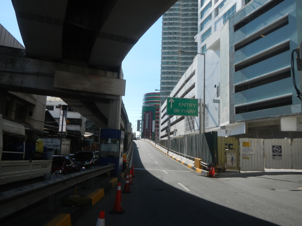 Stadtstraße mit Verkehrskegeln, Fahrzeugen, einer Brücke, Gebäuden mit Schildern, Laternen und Himmel im Hintergrund.