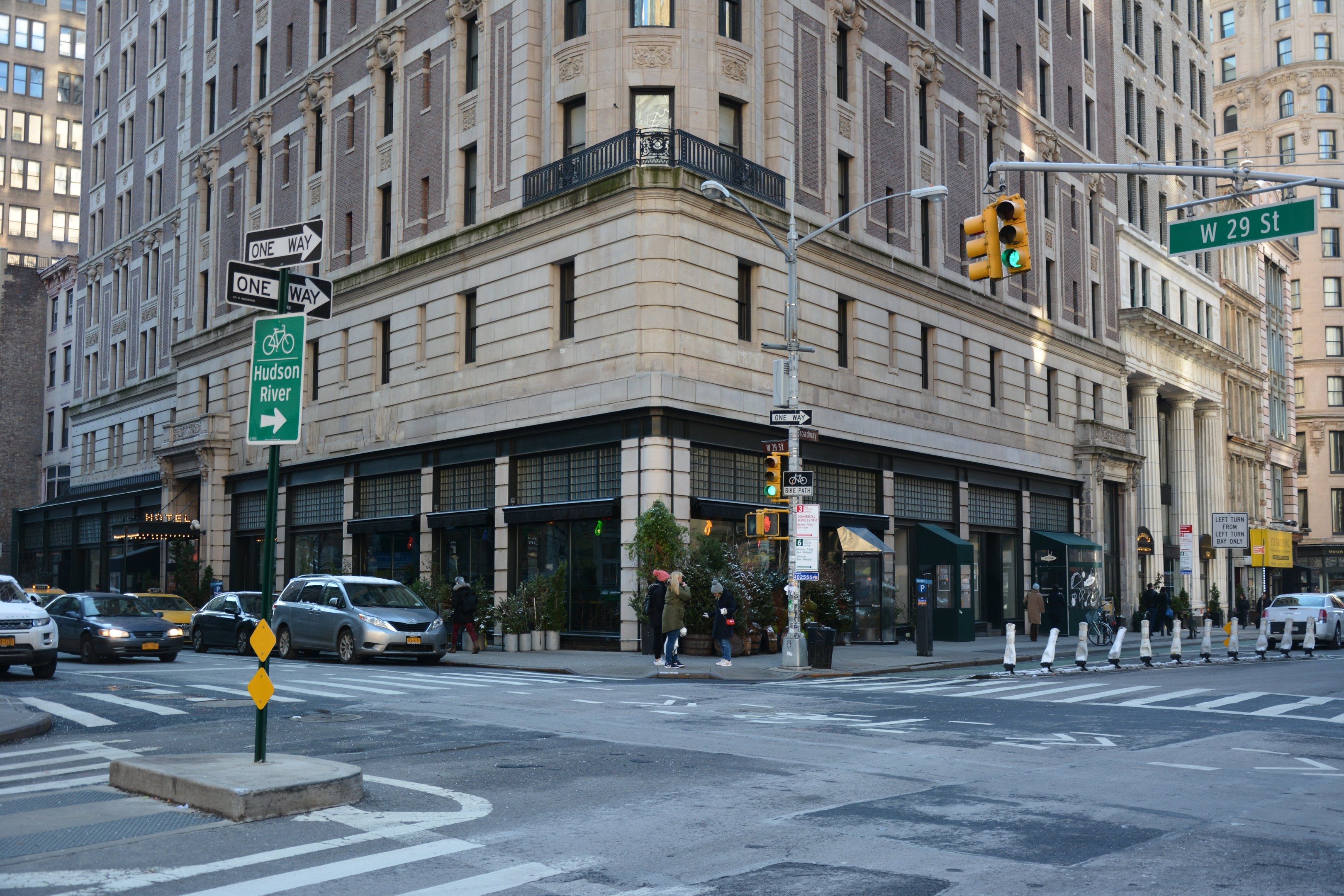 Eine belebte Stadtstraße mit hohen Gebäuden, fahrenden und parkenden Fahrzeugen, Verkehrsampeln, Straßenschildern und Fußgängern auf dem Gehweg.