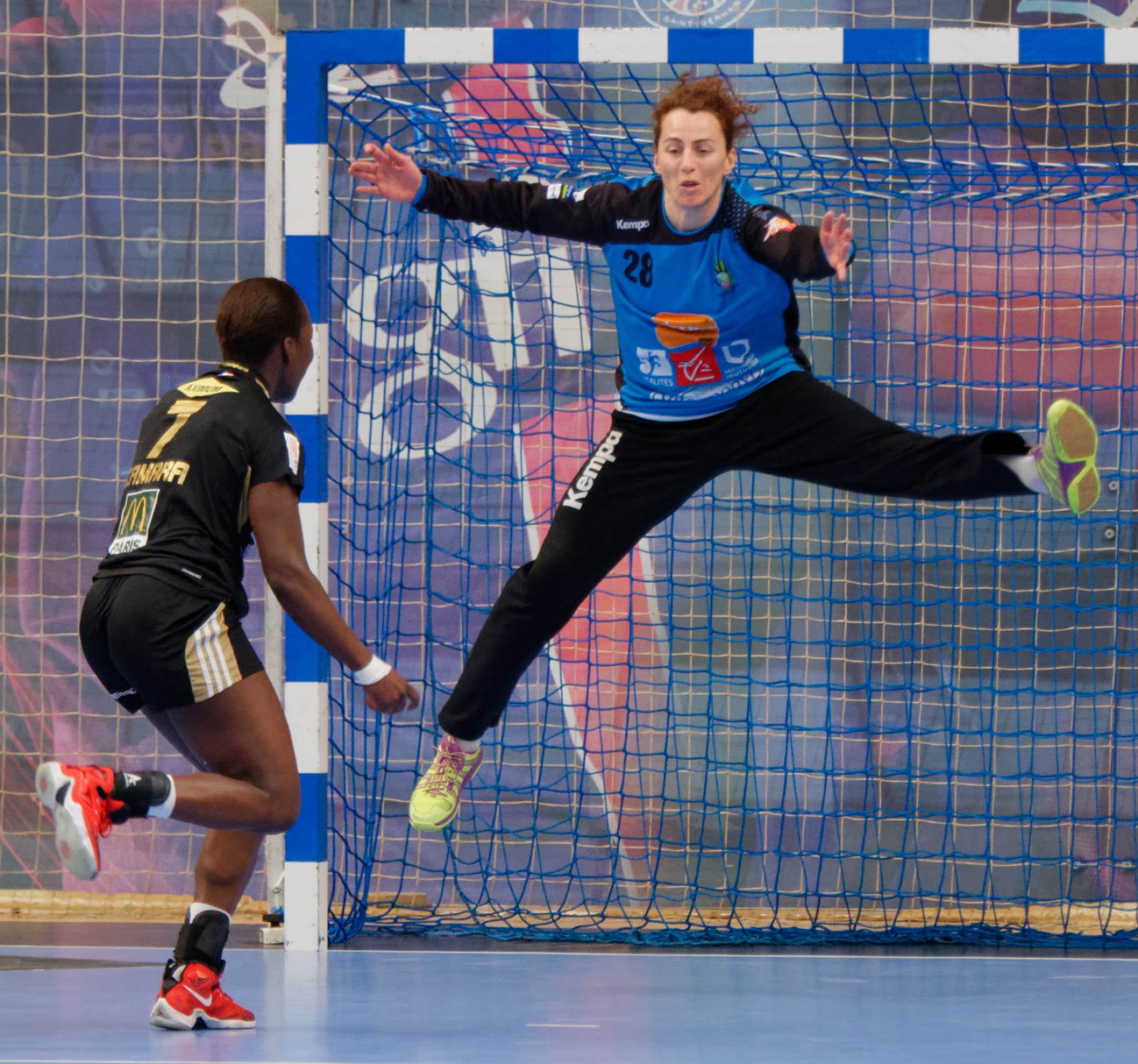 Zwei Frauen beim Handballspielen auf einem Platz mit einem Tor und einer Banner im Hintergrund, beide tragen sportliche Schuhe.