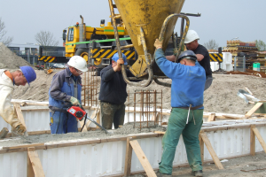 Männer mit Helmen betreiben einen Betonmischer auf einer Baustelle mit Fahrzeugen, Bäumen, Gebäuden und einem klaren Himmel im Hintergrund.