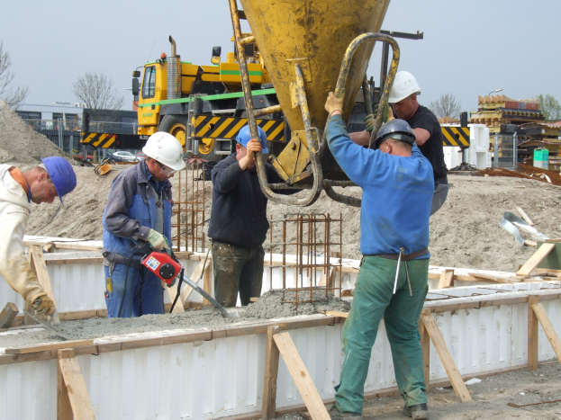 Männer mit Helmen betreiben einen Betonmischer auf einer Baustelle mit Fahrzeugen, Bäumen, Gebäuden und einem klaren Himmel im Hintergrund.