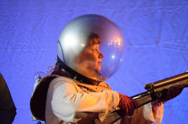 Eine Person in Kleidung und Handschuhen hält eine Waffe, während sie einen Glashelm trägt.