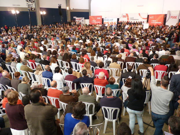 Ein volles Stadion mit sitzenden und stehenden Menschen, eine Bühne mit Personen in der Nähe eines Podiums, zwei sprechende Personen und in der Nähe Banner und Lichter.