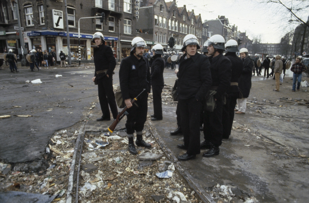 Eine Gruppe von Polizisten in Helmen und mit Waffen in der Hand steht an der Seite einer Straße mit Gebäuden, Schildern, Laternen, Bäumen und einem klaren Himmel im Hintergrund.