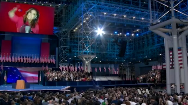 Eine große Menschenmenge sitzt vor einer Bühne mit einem Rednerpult, Mikrofon, Flaggen, einem Bildschirm mit einer Frau und Beleuchtungsausrüstung, wahrscheinlich bei einer Präsidenten-Debatte.