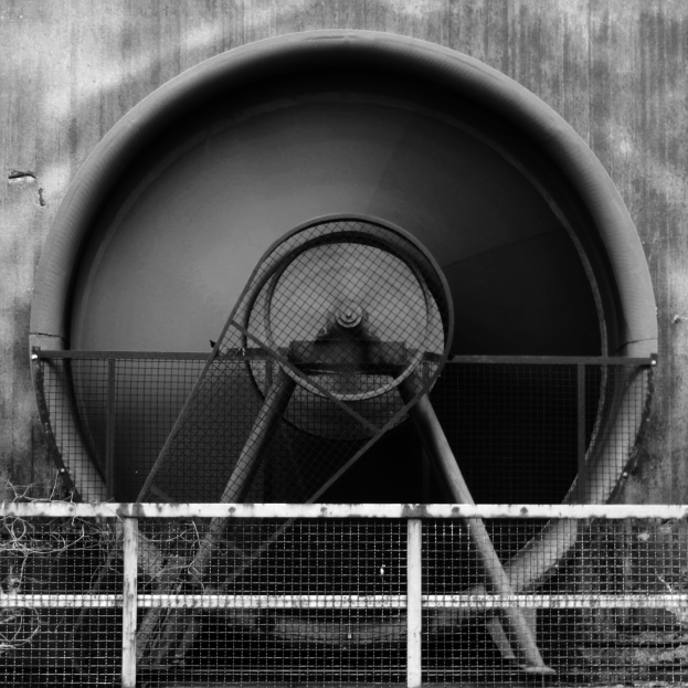 Ein Schwarz-Weiß-Foto eines Abluftgebläses in der Mitte eines Tunnels, umgeben von einem Zaun, mit der Tunnelwand im Hintergrund.