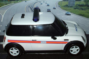 Ein weißer Polizeiwagen ist im Vordergrund geparkt, dahinter fahren mehrere Autos auf der Straße, umgeben von Gras und Bäumen auf beiden Seiten in einer ländlichen Umgebung.