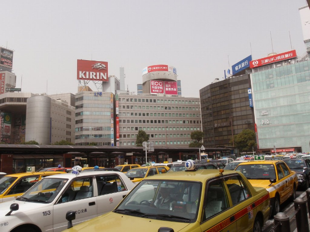 Eine Stadtansicht mit einer Straße mit Fahrzeugen, Gebäuden, Bäumen und Pfählen mit Schildern, die Text anzeigen, einem Zug im Hintergrund und einem Geländer unten rechts.