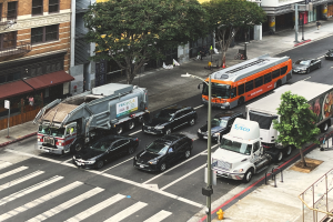 Eine belebte Stadtstraße mit verschiedenen Fahrzeugen, Fußgängern, Bäumen, Laternen, Gebäuden, Geländern, Treppen und Schildern mit Text.