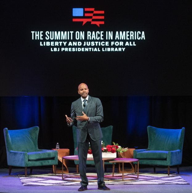 Ein Mann in Blazer, Krawatte und Hemd steht auf einer Bühne und spricht bei "The Summit on Race in America: Liberty and Justice for All"-Veranstaltung in der LBJ Presidential Library. Im Bild sind zwei Stühle, ein Tisch mit Blumenbouquet und einer Flasche darauf, ein Vorhang und ein Bildschirm mit der Aufschrift "The Summit on Race in America: Liberty and Justice for All" zu sehen.