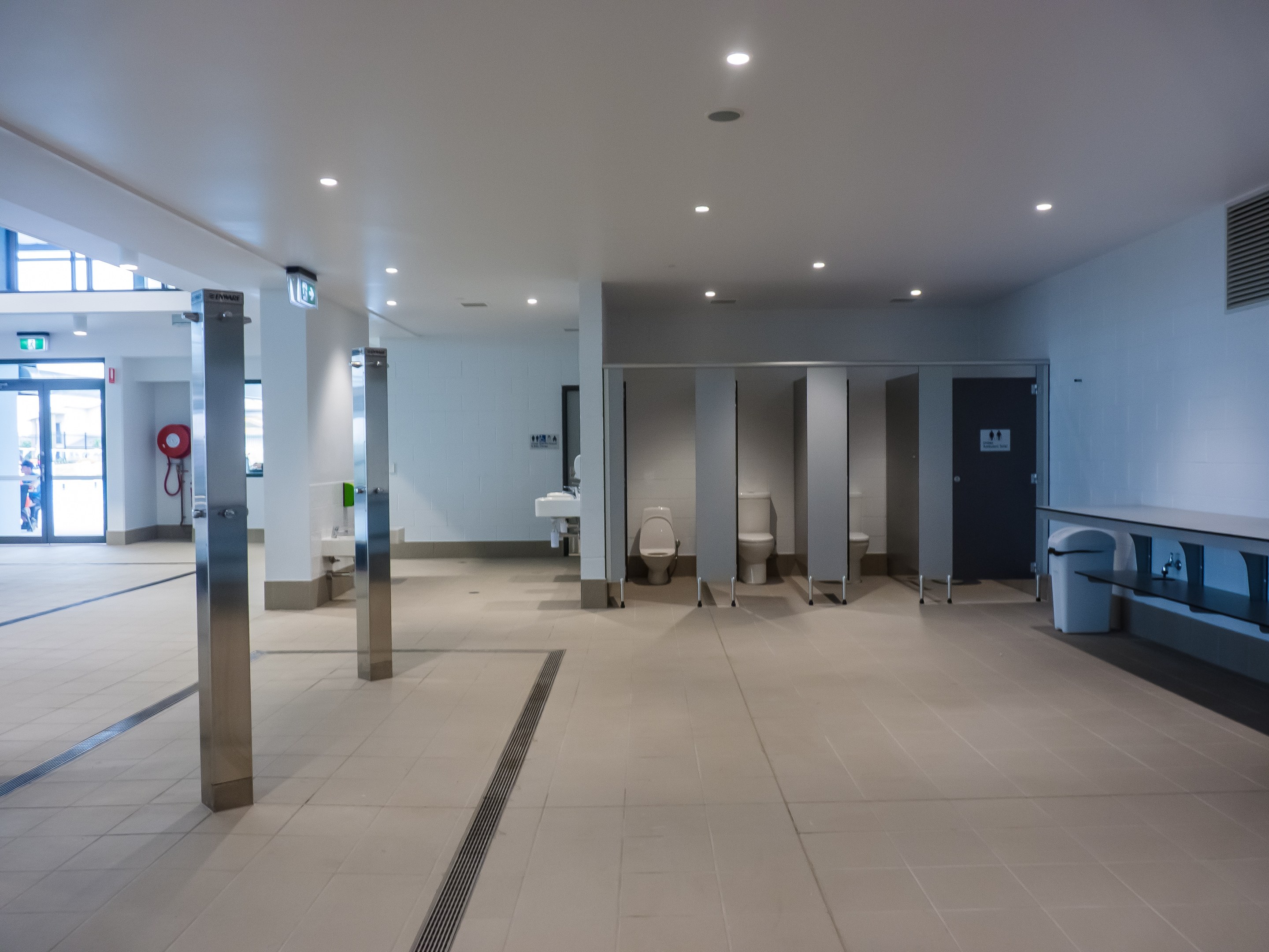 Große öffentliche Toilette mit mehreren Toiletten, tragenden Säulen, einem Mülleimer, einer Tür, einer Tafel, einem Feuerlöscher und Deckenlampen unter einem Dach.