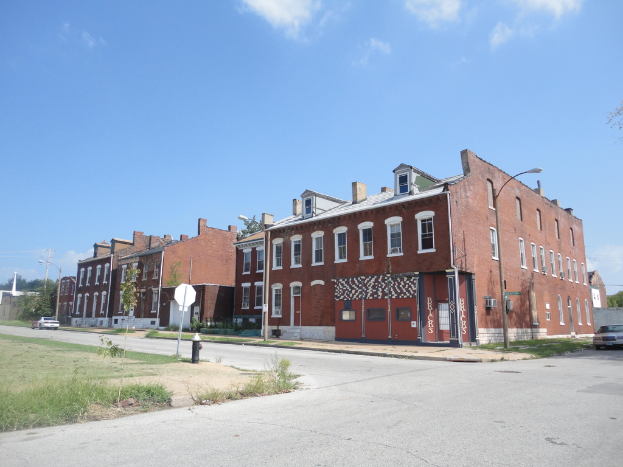 Ein Gebäude mit Masten und Straßenlaternen, umgeben von Bäumen und Fahrzeugen auf beiden Seiten, mit einem Weg, Gras und einem Feuerlöscher unten und Himmel oben.