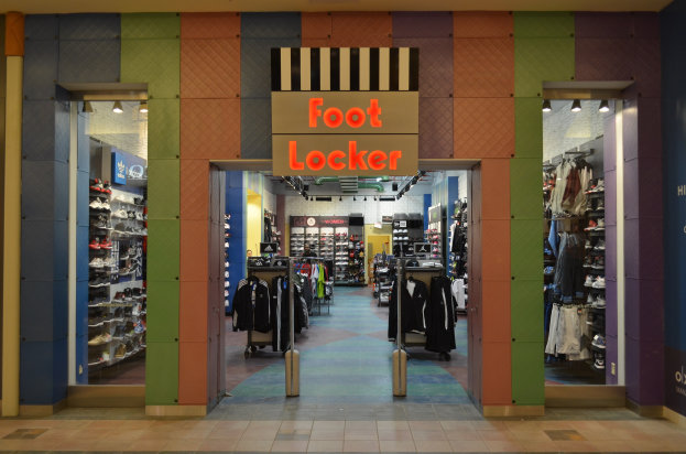 Innenansicht eines Foot Locker-Schuhgeschäfts mit Kleidung an Bügeln und Gestellen unter Deckenbeleuchtung, mit sichtbarem Geschäftsfußboden.