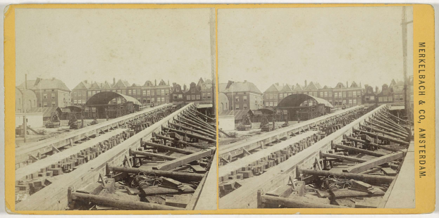 Schwarz-weiß-Foto einer Zugschiene mit Gebäuden im Hintergrund, Text "Merkelbach & Co, Co. Amsterdam" rechts und hölzerne Gegenstände im Vordergrund verstreut.