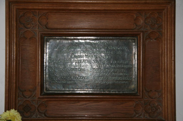 Eine Gedenktafel mit Text wird an einer braunen Wand oder einer Holztür angebracht, mit einem gelben Blumentopf in der linken unteren Ecke.