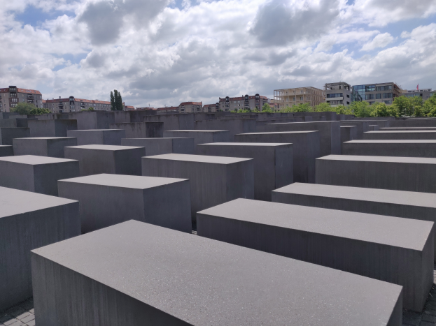 Eine große Anzahl von Betonblöcken in einem Muster angeordnet am Holocaust-Mahnmal für die ermordeten Juden Europas in Berlin, Deutschland, mit Bäumen, Gebäuden und Wolken im Hintergrund.