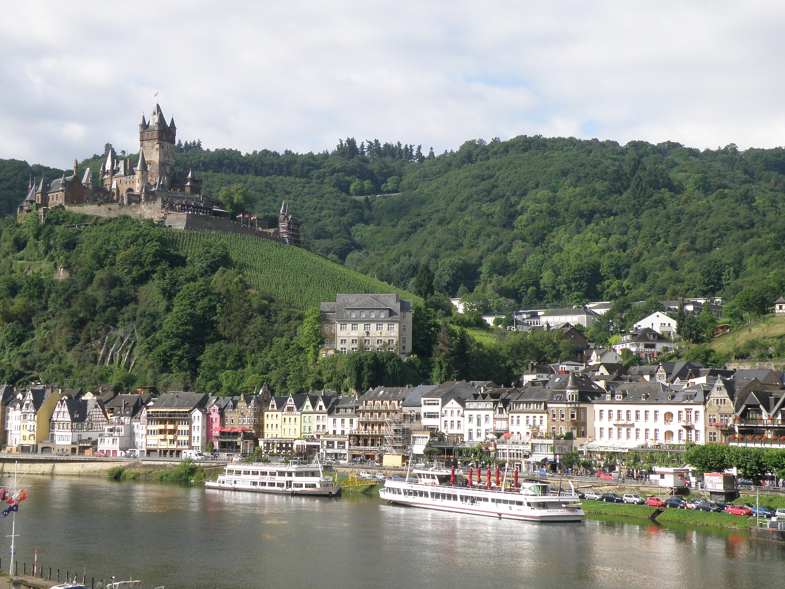 Ein malerischer Blick auf den Rhein in Deutschland mit einer Burg auf einem Hügel, Booten auf dem Wasser, Fahrzeugen auf einer nahen Straße und einer bewölkten Himmel.