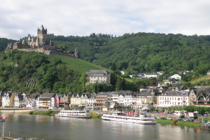 Ein malerischer Blick auf den Rhein in Deutschland mit einer Burg auf einem Hügel, Booten auf dem Wasser, Fahrzeugen auf einer nahen Straße und einer bewölkten Himmel.