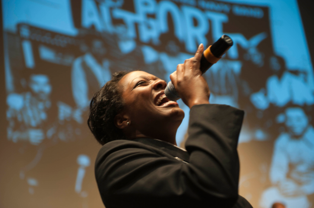 Eine Frau in einem schwarzen Kleid singt in ein Mikrofon auf der Bühne, ihre Augen geschlossen und ihr Gesichtsausdruck zeigt Emotionen, mit ihrem Haar zurückgebunden.