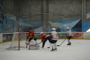 Gruppe von Menschen, die Eis-hockey auf einer Indoor-Eisfläche spielen, mit Helmen und Stöcken, mit einem Tor auf der linken Seite und Bannern im Hintergrund.