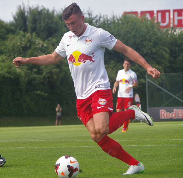 Ein professioneller Fußballspieler, der einen Fußball auf einem grasbewachsenen Feld mit Bäumen und einem klaren blauen Himmel im Hintergrund kickt, mit einem "RB Leipzig"-Schild im Hintergrund.