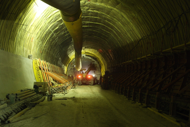 Baustelle mit einem großen Tunnel, Fahrzeugen, verstreuten Holzobjekten, Rohren, einer Wand auf der linken Seite und beleuchteten Lampen im Hintergrund des Tunnels.