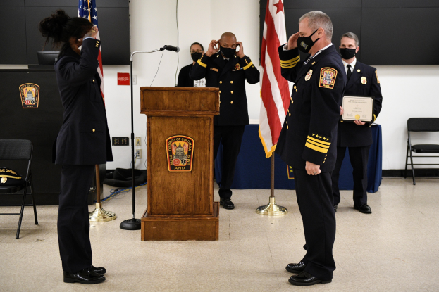 Gruppe uniformierter Personen in Masken an einem Podium mit Mikrofon, das eine Urkunde während einer Vereidigung eines neuen Feuerwehrchefs hält, umgeben von Flaggen, Stühlen, Tischen und einer Wand.