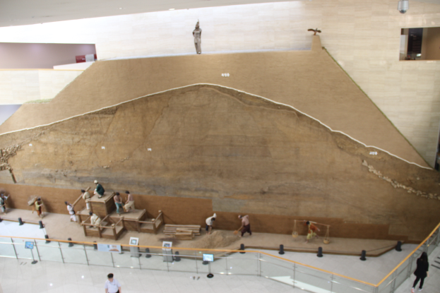 Eine Gruppe von Menschen in einem Museum, umgeben von einem Zaun, Informationsschildern und verschiedenen Gegenständen, mit einer großen Felsformation, wahrscheinlich einem Grabhügel, in der Mitte.