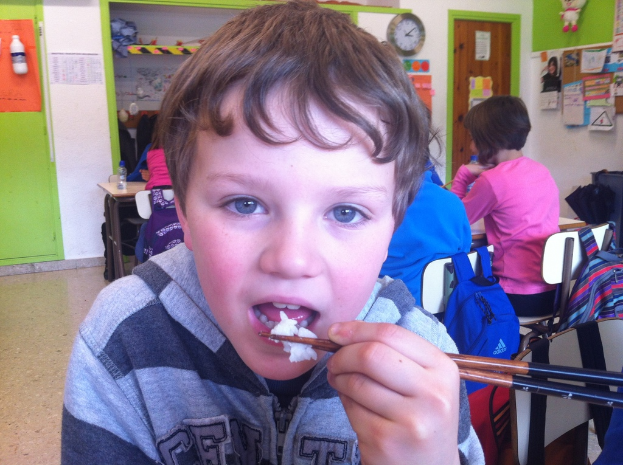 Ein Junge in einer grauen und schwarzen Jacke isst mit Stäbchen in einem Klassenzimmer, mit anderen Kindern, die dahinter sitzen, einer grünen und weißen Wand, Lehrplakaten an einer weißen Wand, einer braunen Tür und einer Wanduhr.