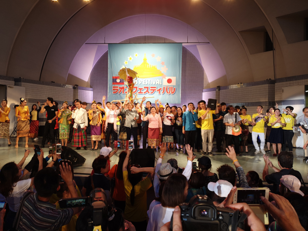 Gruppe von Menschen auf einer Bühne während einer Eröffnungszeremonie, einige halten Kameras oder Telefone und andere Musikinstrumente, mit einem Banner und Deckenlampen im Hintergrund.