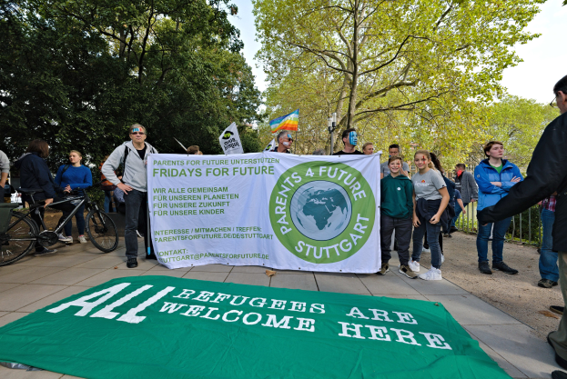 Gruppe von Menschen mit einem Banner "All Refugees Are Welcome Here" und einer Fahne, umgeben von Fahrrädern, einem Zaun, einer Straßenlaterne, einem Schild, Bäumen und einem bewölkten Himmel.