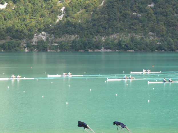 Eine Gruppe von Menschen bei einem Ruderwettbewerb auf einem Gewässer mit Bäumen und einem Hügel im Hintergrund, sowie Kleidung über Stangen am Boden.