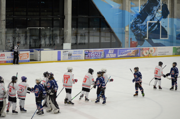 Group of young people playing ice hockey on an indoor rink, wearing helmets and holding sticks, with a person standing on the left side, banners in the background, and glass walls.
