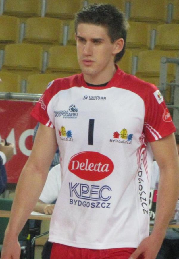 Ein Mann in rot-weißer Handballuniform steht auf einem Feld, umgeben von Zuschauern, mit einem roten Banner im Hintergrund, auf dem "Krasnoyarsk vs Bydgoszcz" steht.