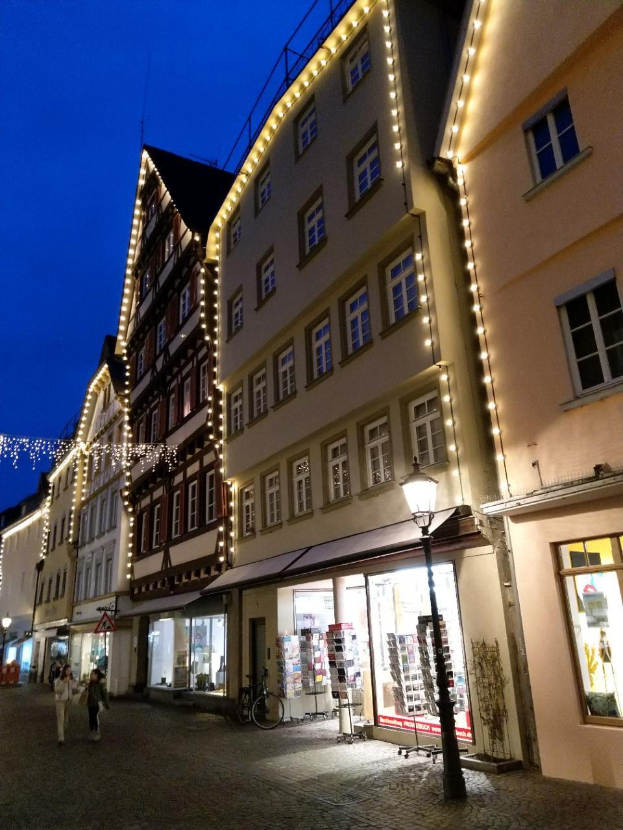 Headsteinstraße in Rothenburg ob der Tauber, Deutschland, geschmückt mit festlichen Weihnachtslichtern, mit Menschen, die spazieren gehen und ein Fahrrad in der Nähe geparkt ist, flankiert von Gebäuden mit Fenstern unter einem sichtbaren Himmel.