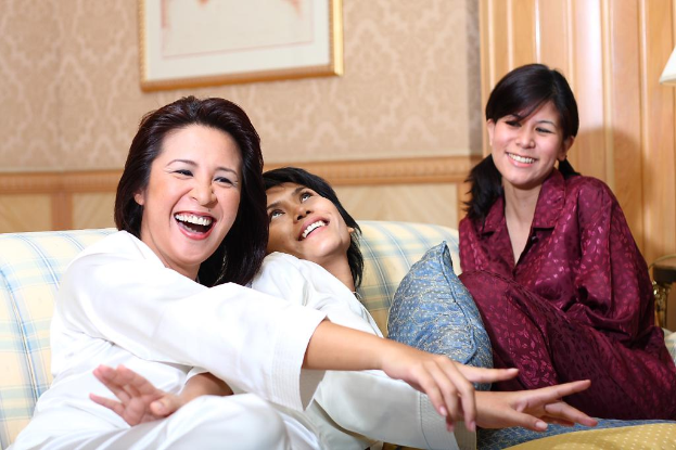 Drei Frauen lachen zusammen, mit einem Kissen neben der mittleren Frau und einem gerahmten Bild im Hintergrund.