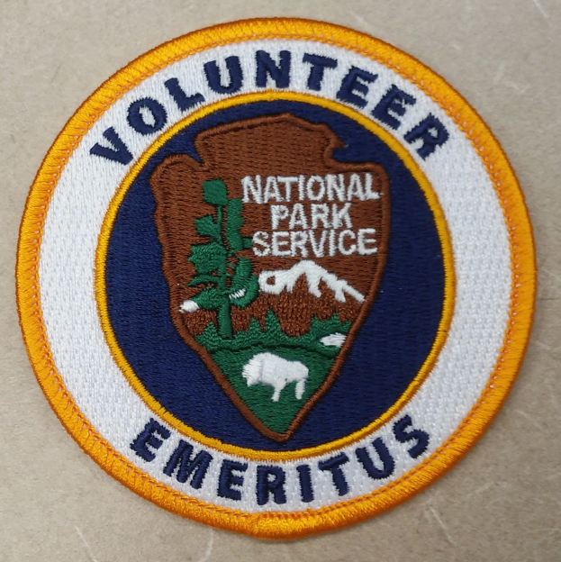 Ein kreisförmiges Patch mit einem blauen Rand, das die fette schwarze Schrift "Volunteer National Park Service Emeritus" in der Mitte eines gelben Kreises auf einem weißen Hintergrund zeigt.