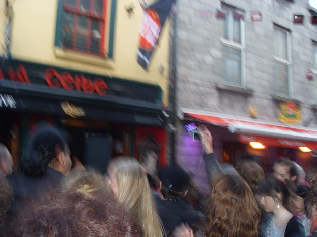 Ein unscharfer Bildausschnitt mit Menschen auf der Straße unten und Gebäuden, Lichtern, Fenstern und einer Fahne oben.