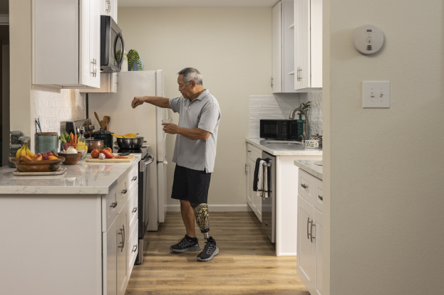 Ein Mann steht in einer Küche mit weißen Schränken und hält ein Glas in der Hand, während verschiedene Küchenutensilien, einschließlich Obst und Besteck, auf einer Arbeitsplatte links von ihm liegen.