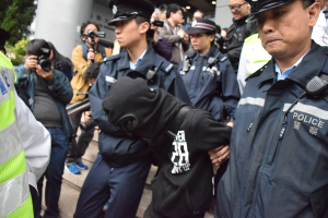 Eine Gruppe von Polizeibeamten steht vor einer Menschenmenge, einige tragen Mützen und halten Kameras, im Hintergrund ein Gebäude mit Säulen und Lampen und links Bäume.