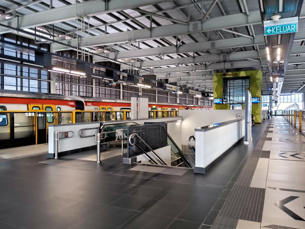 Ein Bahnhof mit einem Zug auf den Schienen, eine Plattform mit Geländern, Treppen mit Handläufen, Schilder mit Text, Lampen an der Decke und Glaswände.
