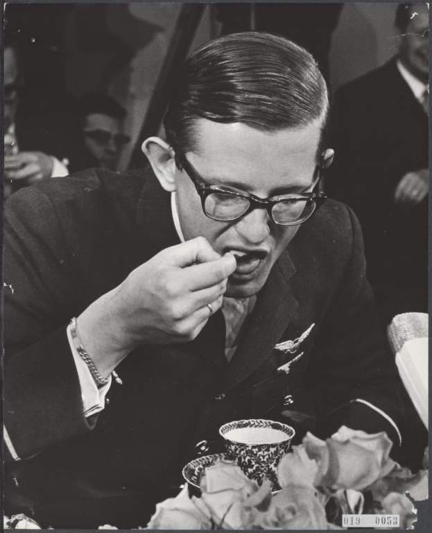 Ein Mann in einem Blazer und mit Brille sitzt an einem Tisch mit einer Tasse und Untertasse, isst, mit ein paar anderen Menschen im schwarz-weißen Hintergrund.